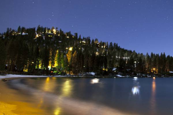 Coast of Lake Tahoe at night in front of a forested hill with lights from buildings.